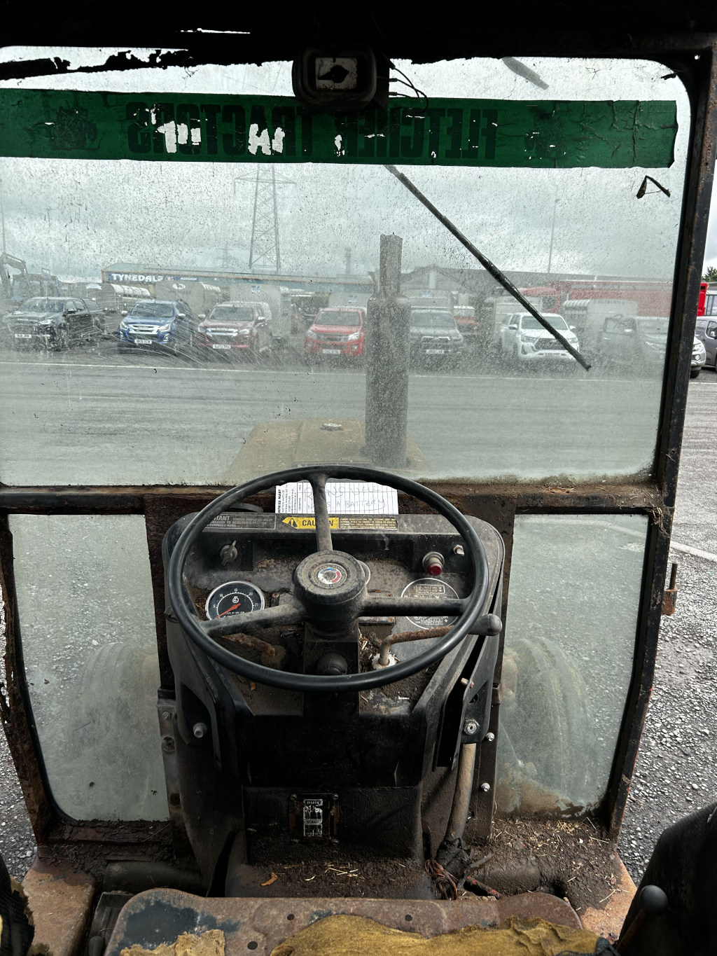 Interior of an old, rusty tractor cab. The view from the driver's seat looks out onto a parking lot with several cars visible through a dirty and dusty windshield. A green warning sticker is visible across the top of the windshield. The tractor's steering wheel, dashboard, and controls are worn and show significant signs of age and use.