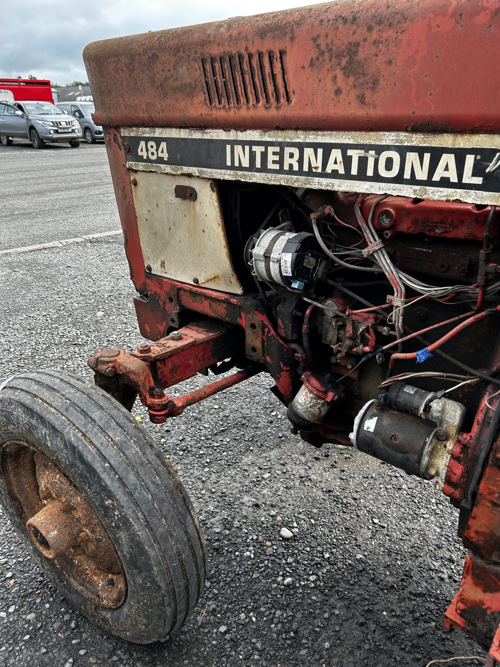 Close-up view of the front of an old, rusty International Harvester 484 tractor. The tractor shows significant wear and tear, with rust visible on the body and exposed mechanical parts. The focus is on the engine compartment and the front wheel. The background shows a parking lot with other vehicles.