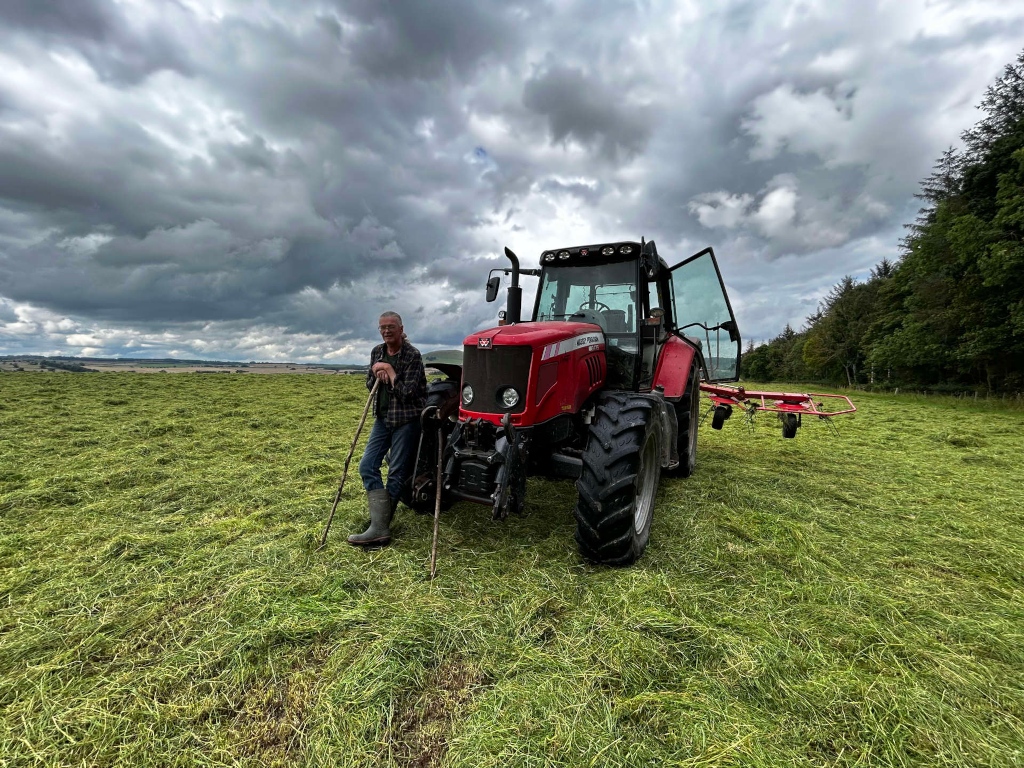 Charlie standing proudly beside his red tractor in a freshly cut hay field. The sky is dramatic and cloudy, adding a sense of atmosphere to the scene. He is holding two walking sticks, suggesting a rural lifestyle and perhaps hinting at the hard work and tradition involved in farming. The overall impression is one of peaceful rural life, possibly reflecting the satisfaction of a job well done.