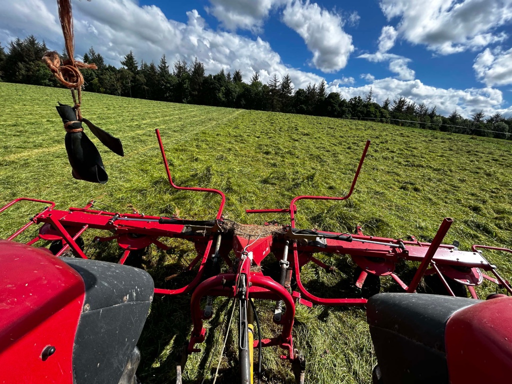 Rear view from the driver's seat of a red tractor with a hay tedder attached.  The tedder is working in a freshly cut field of green grass, under a bright, partly cloudy sky. A wooded area is visible in the background. A dark object hangs from a rope above the tractor.