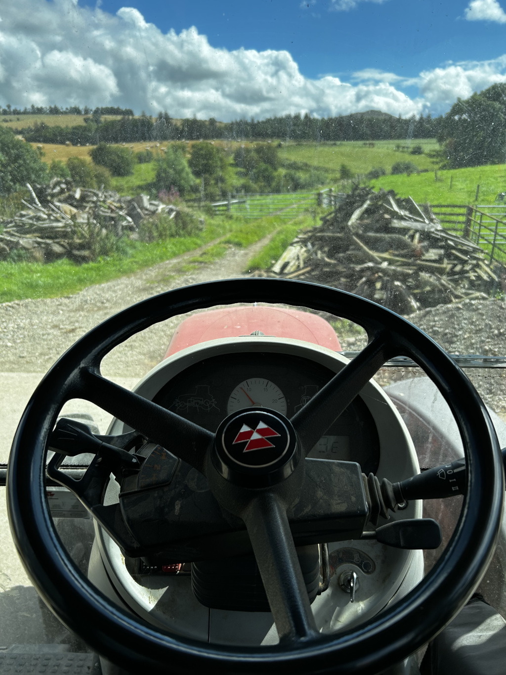 View from inside a tractor, specifically the steering wheel and dashboard. The foreground is dominated by the tractor's steering wheel, which displays the Massey Ferguson logo. The background shows a rural landscape, including fields, piles of lumber, and a partly cloudy sky.