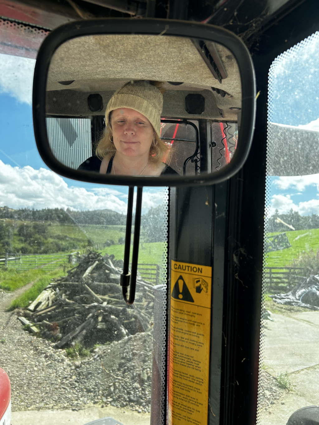 Reflection in the side mirror of a tractor. The reflection shows Leonie with blonde hair wearing a light beige beanie, sitting in the driver's seat. The background of the reflection shows a rural landscape with a pile of wood and a field. A caution sticker is visible on the tractor's frame. The overall impression is of a woman working on a farm or in a rural setting.