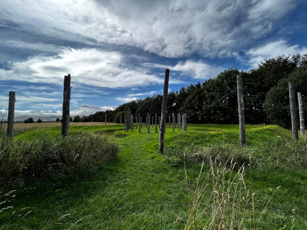 Grassy field with a line of weathered wooden posts extending into the middle distance. The posts are irregularly spaced and appear to be remnants of a structure or possibly a historical marker. Behind the posts is a line of trees, and a partly cloudy sky is visible above. The overall impression is one of a serene, somewhat desolate, yet historically significant landscape.