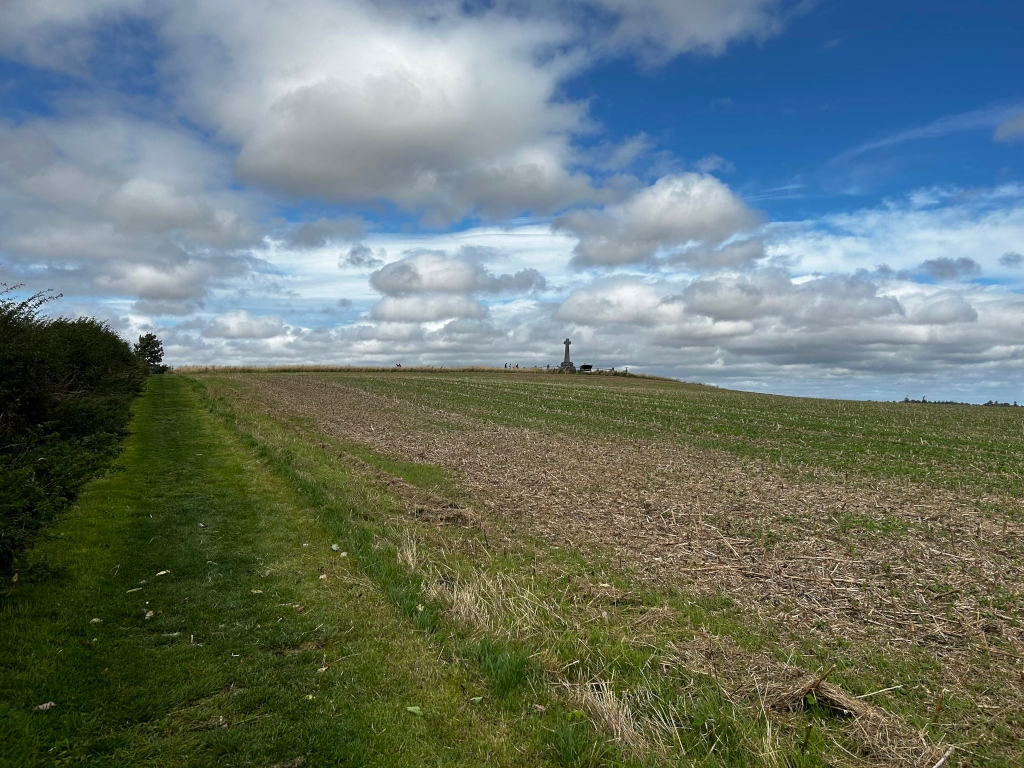 Grassy path bordering a harvested field that stretches towards a distant hilltop. Atop the hill is a simple stone monument or cross, possibly a war memorial. The sky is partly cloudy, with fluffy white clouds against a clear blue backdrop. The overall impression is one of peaceful rural landscape, marked by a sense of remembrance suggested by the monument.
