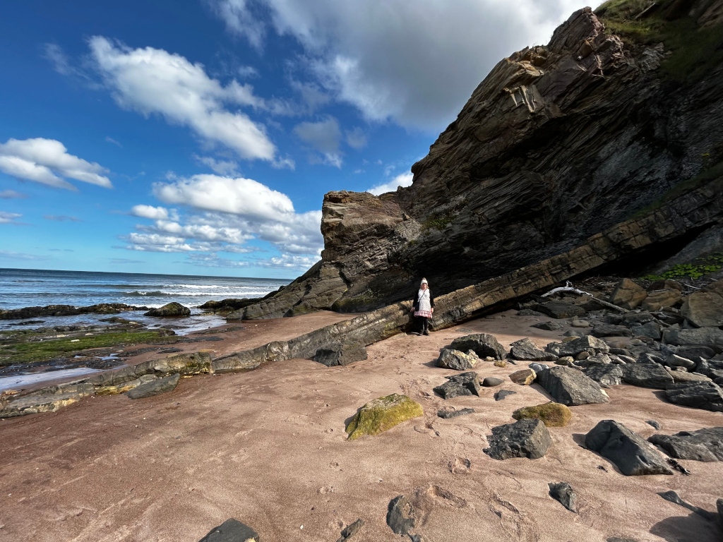 Leonie standing on a sandy beach next to a large, dark-coloured rock formation. The rock face is steeply sloped and shows distinct layers of rock, suggesting geological stratification. The beach is mostly sandy, with scattered dark rocks and patches of greenish-brown algae visible near the water's edge. The ocean is visible in the background, with relatively calm waves. The sky is mostly clear and blue, with some fluffy white clouds. The overall scene is one of a dramatic coastal landscape.