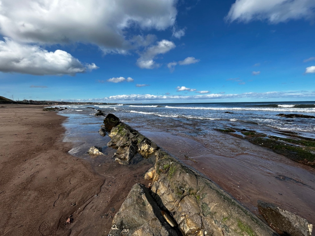 Sandy beach with dark rocks extending from the shore into the shallow ocean water. The ocean is relatively calm with small waves. The sky is mostly sunny with fluffy white clouds. The overall scene is peaceful and evocative of a coastal landscape.