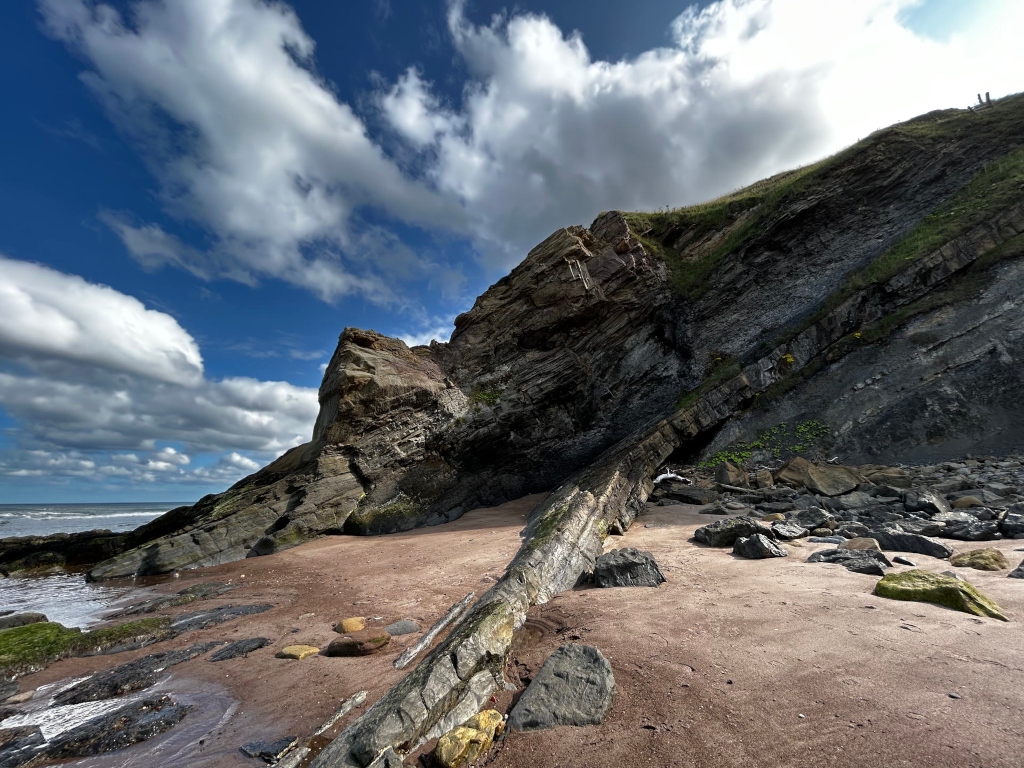 Dramatic coastal scene. A large, layered rock formation dominates the frame, its dark, striated surfaces contrasting sharply with the lighter-coloured sand below. A long, dark log or piece of driftwood lies diagonally across the foreground sand, leading the eye towards the base of the cliff. The sky above is a vibrant blue, punctuated by fluffy white clouds that add depth and texture to the scene. The ocean is visible in the lower left, its waves gently lapping at the shore. The color palette ranges from the deep blues and grays of the rock and sea to the warm browns and tans of the sand, creating a visually striking composition.