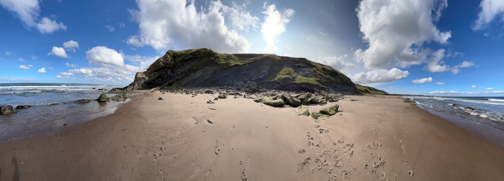 Panoramic view of a sandy beach with a low cliff in the background.  The beach is relatively empty except for some rocks and animal tracks in the sand. The sea is calm with small waves, and the sky is mostly sunny with scattered clouds.