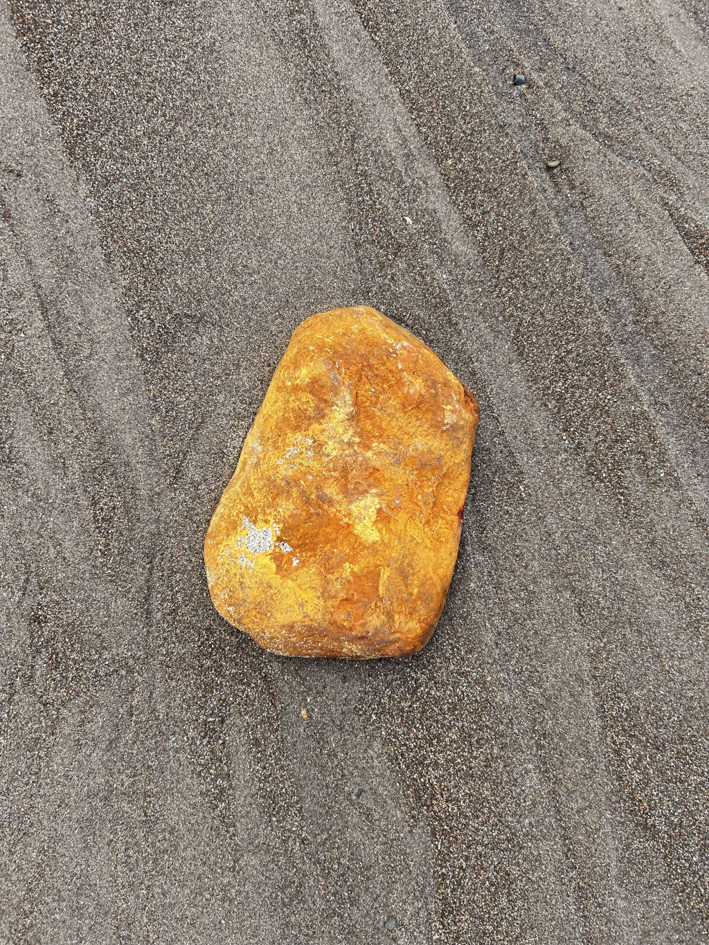 Single, roughly oval-shaped, yellow-gold rock resting on a bed of dark gray sand. The sand shows subtle patterns suggesting recent movement, perhaps from wind or water. The contrast between the rock's colour and the sand is striking.