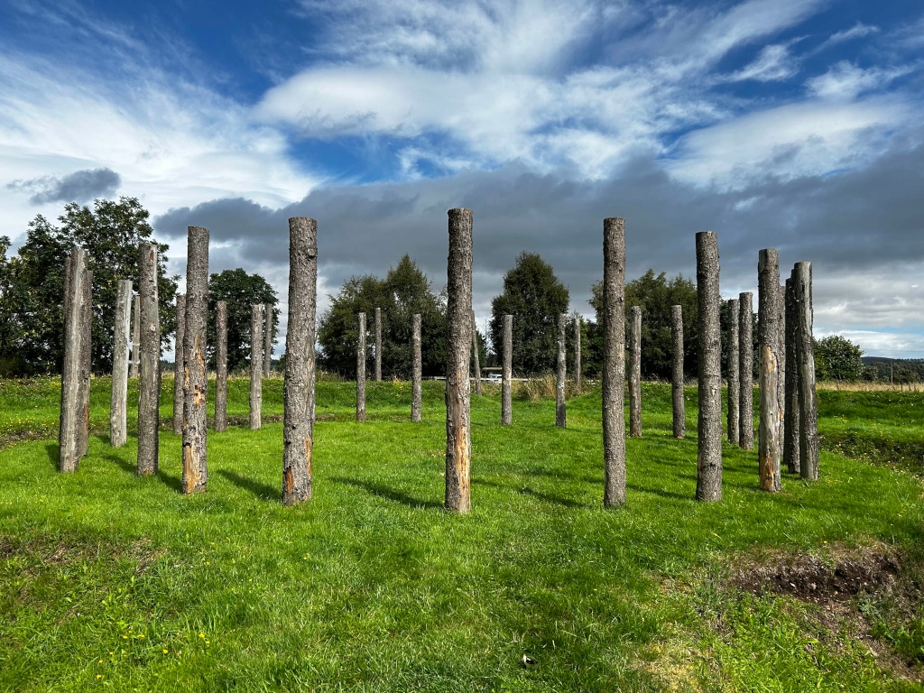 Field of short, bright green grass under a partly cloudy sky. Arranged in a loose circle within the field are numerous upright, weathered grey-brown wooden posts or poles of varying heights. The scene is peaceful and evokes a sense of quiet contemplation or perhaps a forgotten ritual site.
