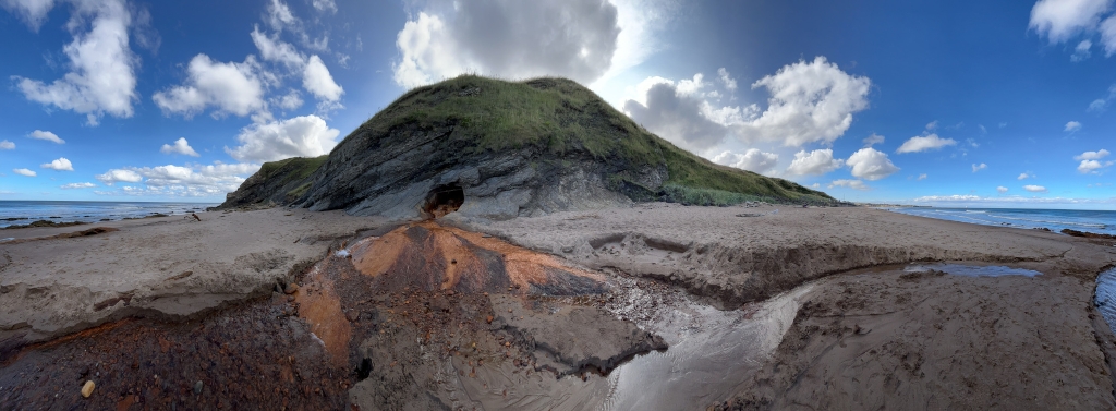 Panoramic view of a sandy beach with a small, grassy hill in the background.  A cave-like opening is visible near the base of the hill, from which a stream of reddish-brown water flows down onto the beach, creating a distinctive pattern in the sand. The sky is mostly clear with scattered clouds, and the ocean extends to the horizon.