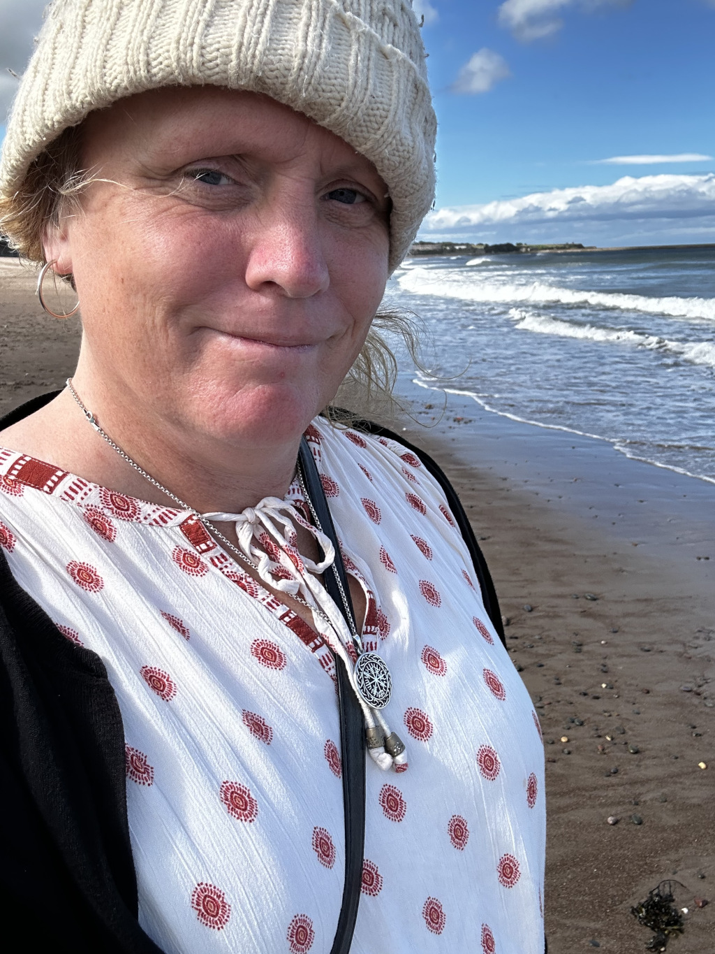Close-up selfie of a fair-skinned woman named Leonie wearing a cream-coloured knit beanie and a white blouse with a red pattern. She's wearing a silver necklace with a circular pendant and has hoop earrings. The background is a blurred-out beach scene with the ocean and a partly cloudy sky. The woman has a somewhat serious yet calm expression.
