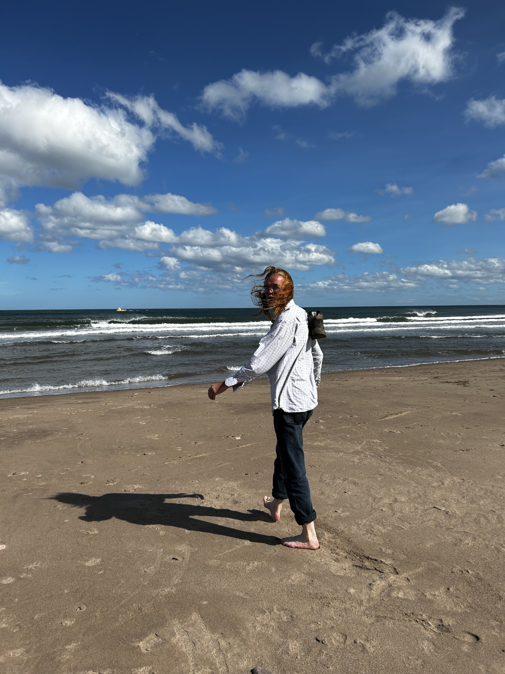 Charlie with long, red hair walking barefoot on a sandy beach towards the camera. Their hair is blowing in the wind, and they are carrying a shoe or boot on their shoulder. The background features a calm ocean under a bright blue sky with fluffy white clouds. The overall impression is one of freedom and casual enjoyment of a sunny day at the beach.