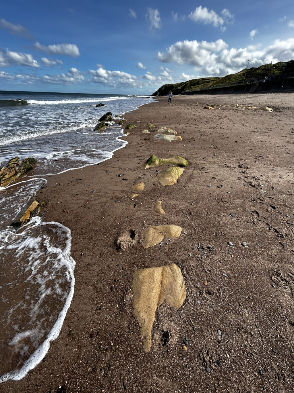 Sandy beach with a person walking in the distance. The foreground features a series of large, light-coloured rocks partially embedded in the dark sand, close to the water's edge where small waves are breaking. The sky is bright blue with fluffy white clouds. The overall impression is one of a peaceful, sunny day at the coast with a solitary figure.