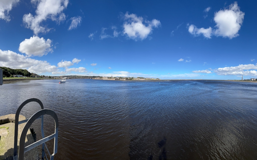 Panoramic view of a wide river or estuary on a sunny day.  A sailboat is visible in the mid-ground, and a town or city is seen in the far distance across the water. The sky is mostly blue with fluffy white clouds. In the foreground, a metal ladder attached to a concrete structure is partially visible. The overall feel is serene and peaceful.