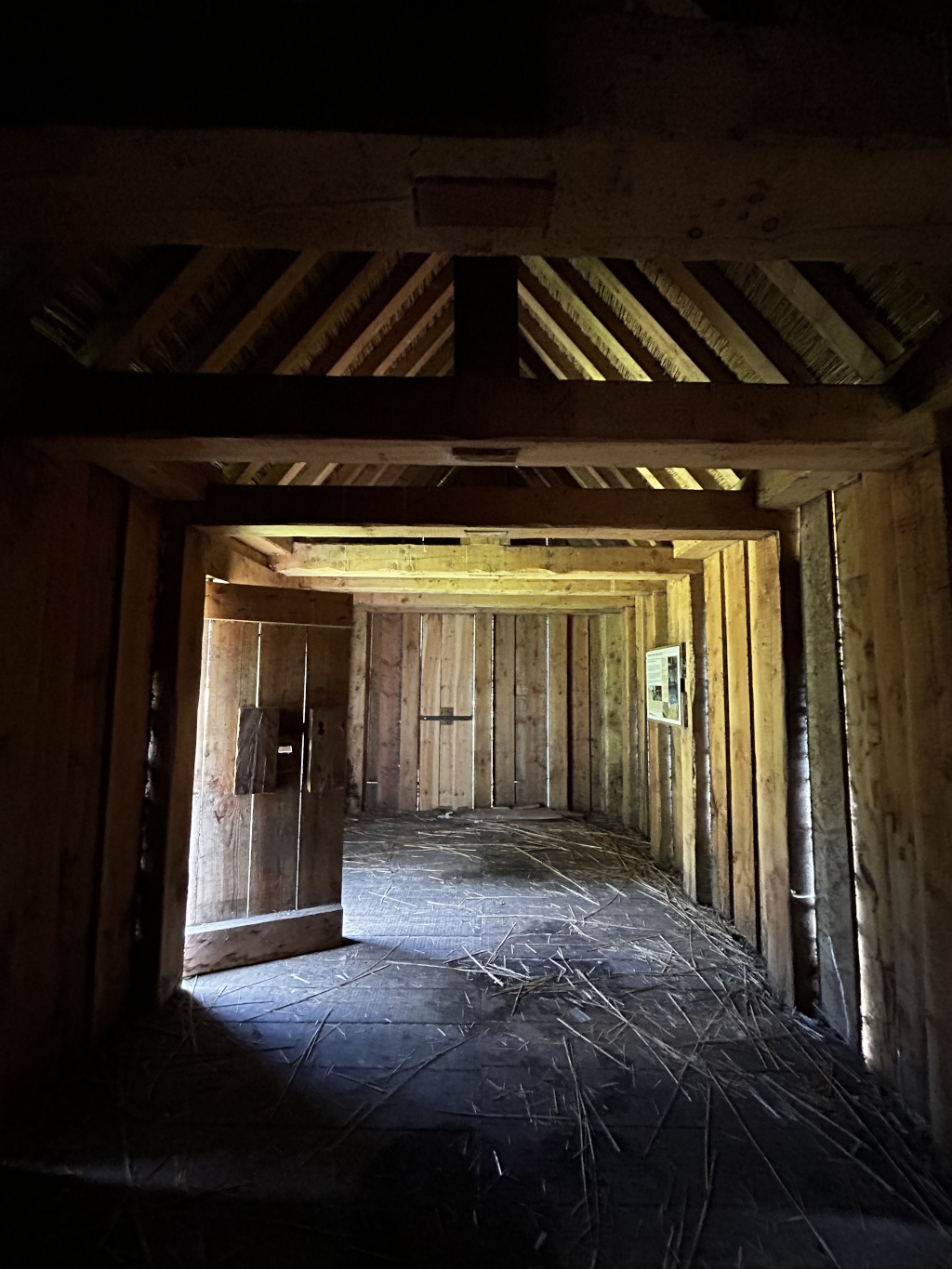The interior of a long, narrow, rustic wooden structure, possibly a reconstructed historical building or a barn. The structure is made of vertical wooden planks, with a thatched roof supported by heavy wooden beams. A single wooden door is ajar, letting in light that illuminates the dusty floor strewn with straw or hay. The overall atmosphere is dark, secluded, and somewhat mysterious.