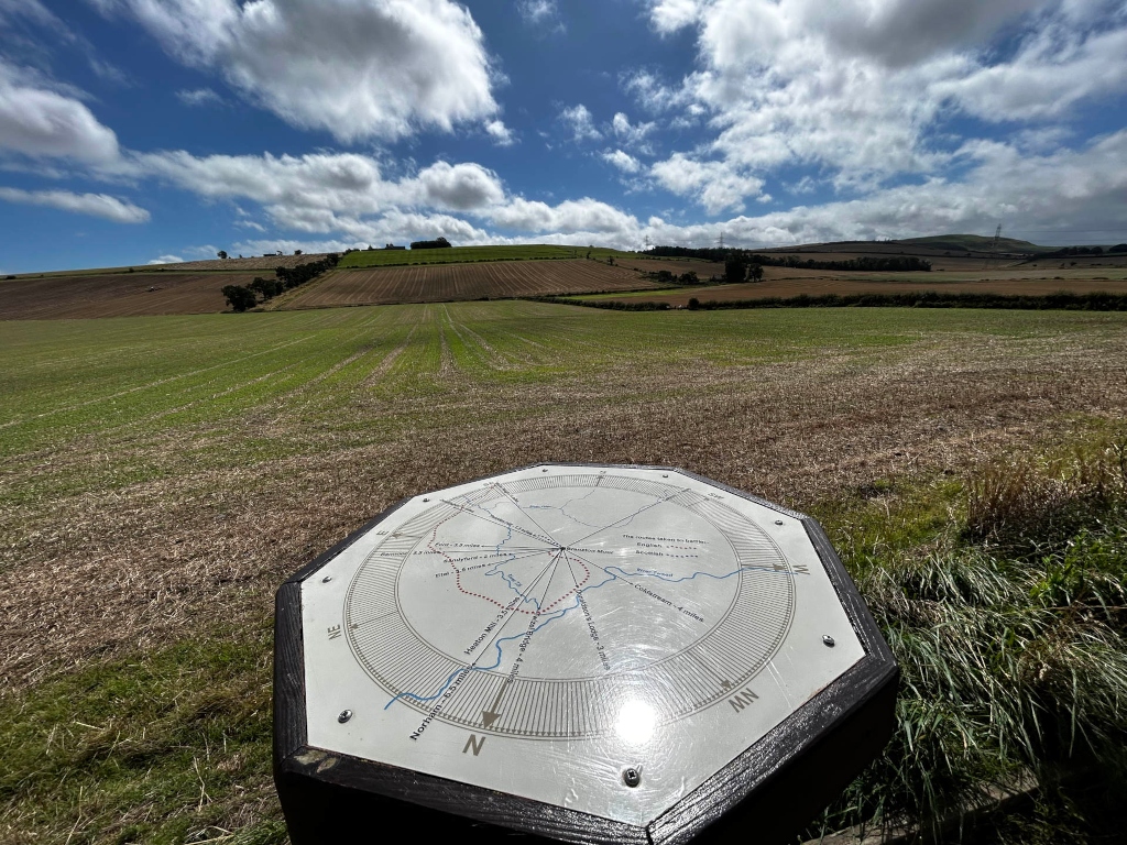 Ground-level view of a countryside scene dominated by expansive agricultural fields under a partly cloudy sky. In the foreground, a prominent octagonal sign, resembling a compass or waymarker, displays a simplified map of the surrounding area with directional markings and distances to various points of interest. The map is printed on a light-coloured surface and fixed to a dark-colored wooden base. The background reveals rolling hills with patches of harvested and growing crops, suggesting a rural setting. 