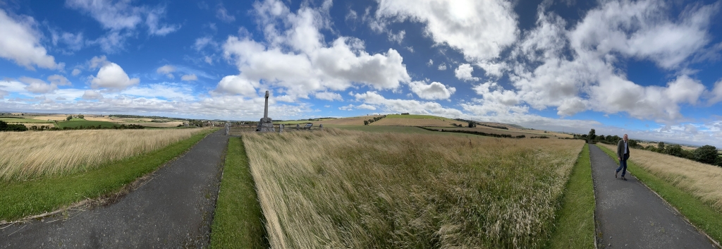 Panoramic view of a rural landscape, likely in the British Isles, under a partly cloudy blue sky. The foreground features two paved pathways flanking a field of tall, golden-brown grass. In the middle ground, a stone war memorial is visible, suggesting a site of historical significance. Charlie walks along one of the paths in the distance. The background shows gently rolling hills and fields, indicating a tranquil and expansive countryside setting. The overall impression is one of serenity and quiet remembrance.