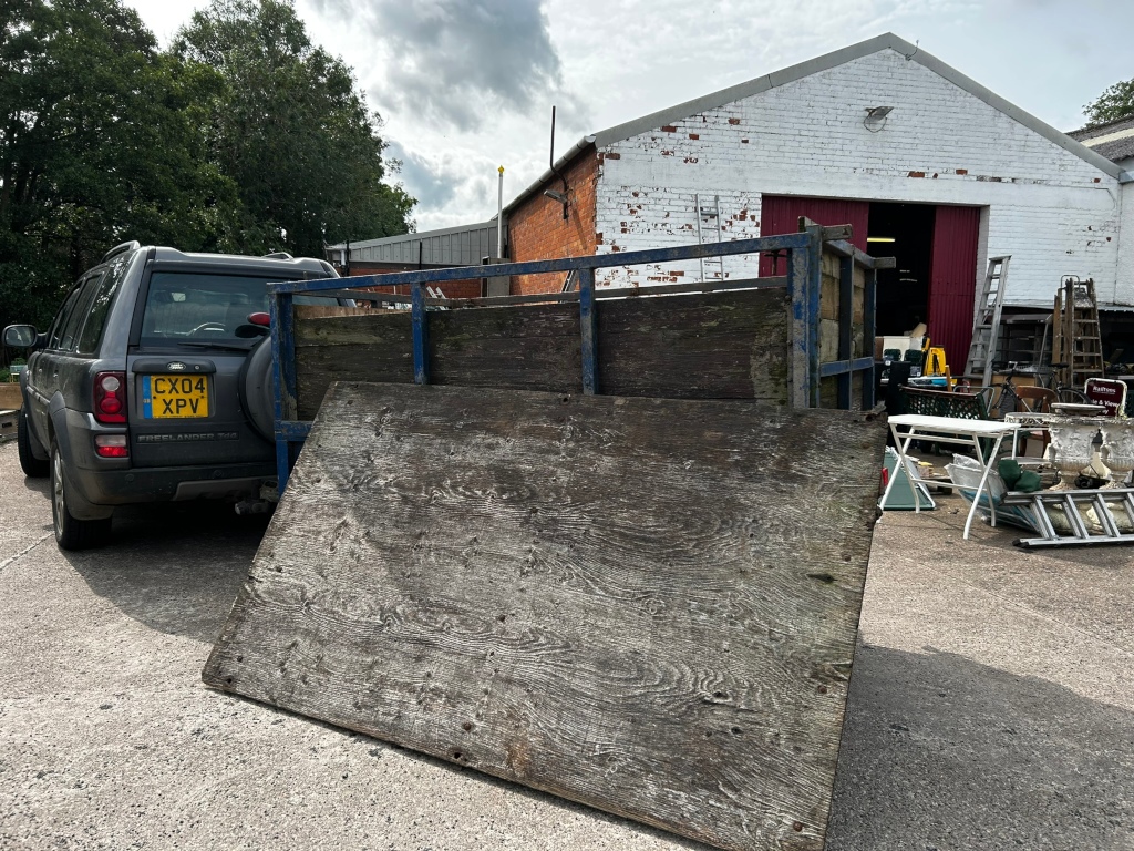 Land Rover Freelander parked in front of a white brick building. The Land Rover is connected to a trailer carrying a large, weathered piece of plywood. The background is cluttered with various items, suggesting a yard sale, reclamation yard, or similar setting. The overall impression is one of casual, everyday activity.