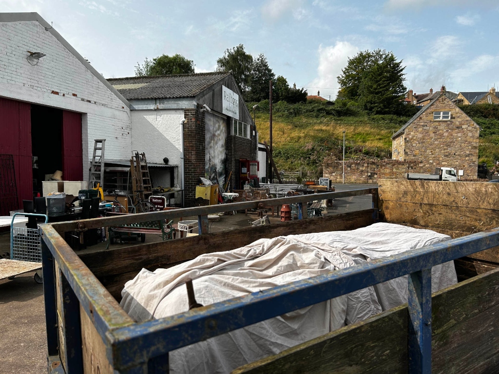 Back of a cluttered salvage yard or reclamation business. A flatbed truck is in the foreground, partially covered with a tarp. The background shows various buildings, some appearing to be industrial or storage units, and a stone building possibly residential. Various items, including ladders, furniture, and other salvage materials, are scattered around the yard. The overall feel is one of organised chaos, suggesting a business focused on recycling and repurposing materials.