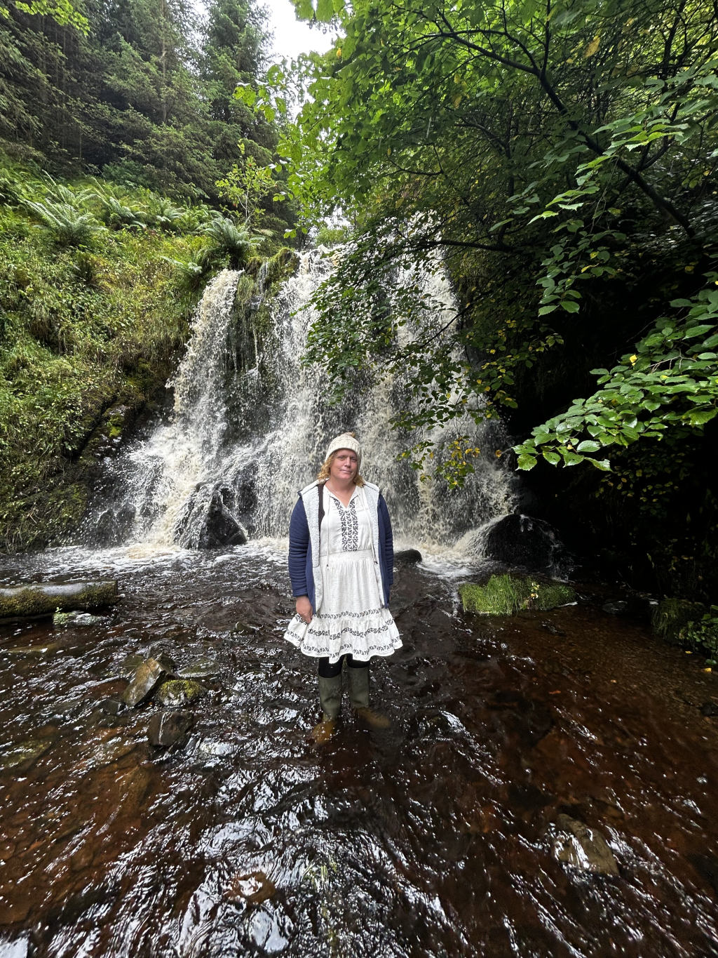 Leonie standing in a shallow stream in front of a waterfall. She's wearing a white dress, a dark blue cardigan, and a light-colored knit hat. The setting is lush and green, with abundant foliage surrounding the waterfall. The water in the stream appears dark and somewhat muddy. The overall mood is tranquil and natural.