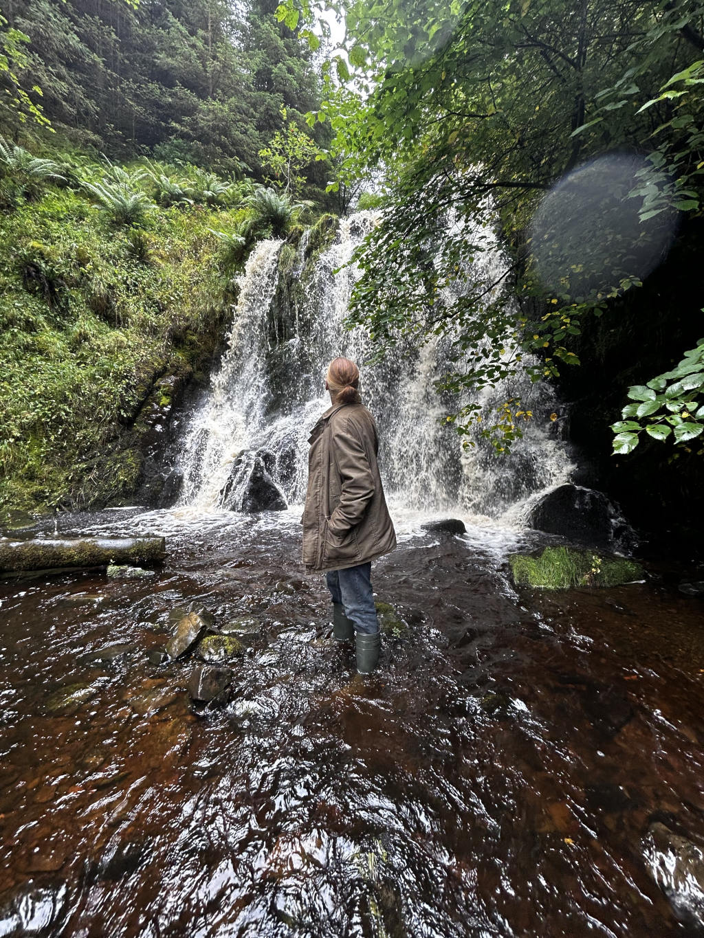 Charlie standing in a shallow stream at the base of a waterfall in a lush, green forest. Charlie, whose back is to the camera, seems to be observing the waterfall and the surrounding natural environment. The overall mood is serene and contemplative, emphasising the beauty of the natural landscape.