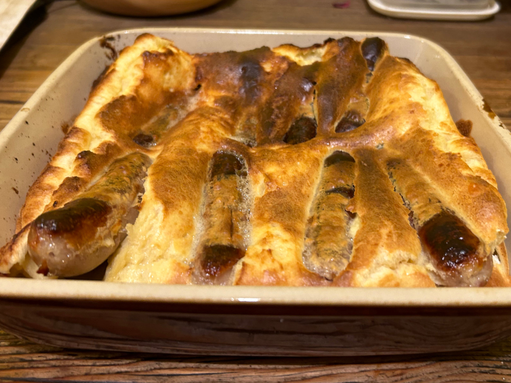 Toad in the hole, a savoury British dish. It consists of sausages baked in Yorkshire pudding batter, resulting in a golden-brown, puffed-up batter surrounding the sausages. The dish is presented in a rectangular baking dish.