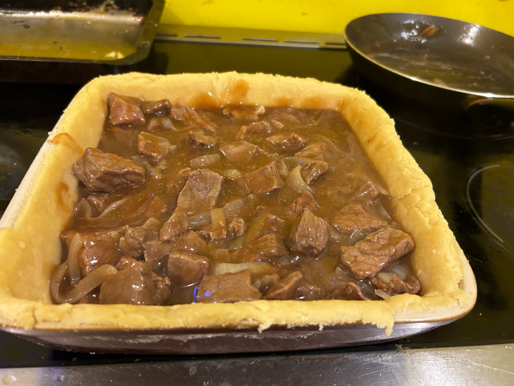 Partially-assembled steak and onion pie. A pastry crust is in a baking dish, filled with a rich, brown gravy containing chunks of steak and onions. Other baking pans are visible in the background, suggesting a cooking process in progress.