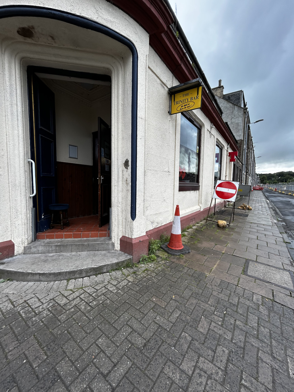Exterior of a pub called The Trinity Bar in what appears to be a small town or village.  The pub's entrance is partially open, revealing a glimpse of the interior. A No Entry sign is present on the sidewalk, suggesting potential roadworks or closures nearby. A traffic cone sits on the pavement near the pub's entrance. The overall atmosphere feels quiet and somewhat deserted, with overcast skies visible in the background.