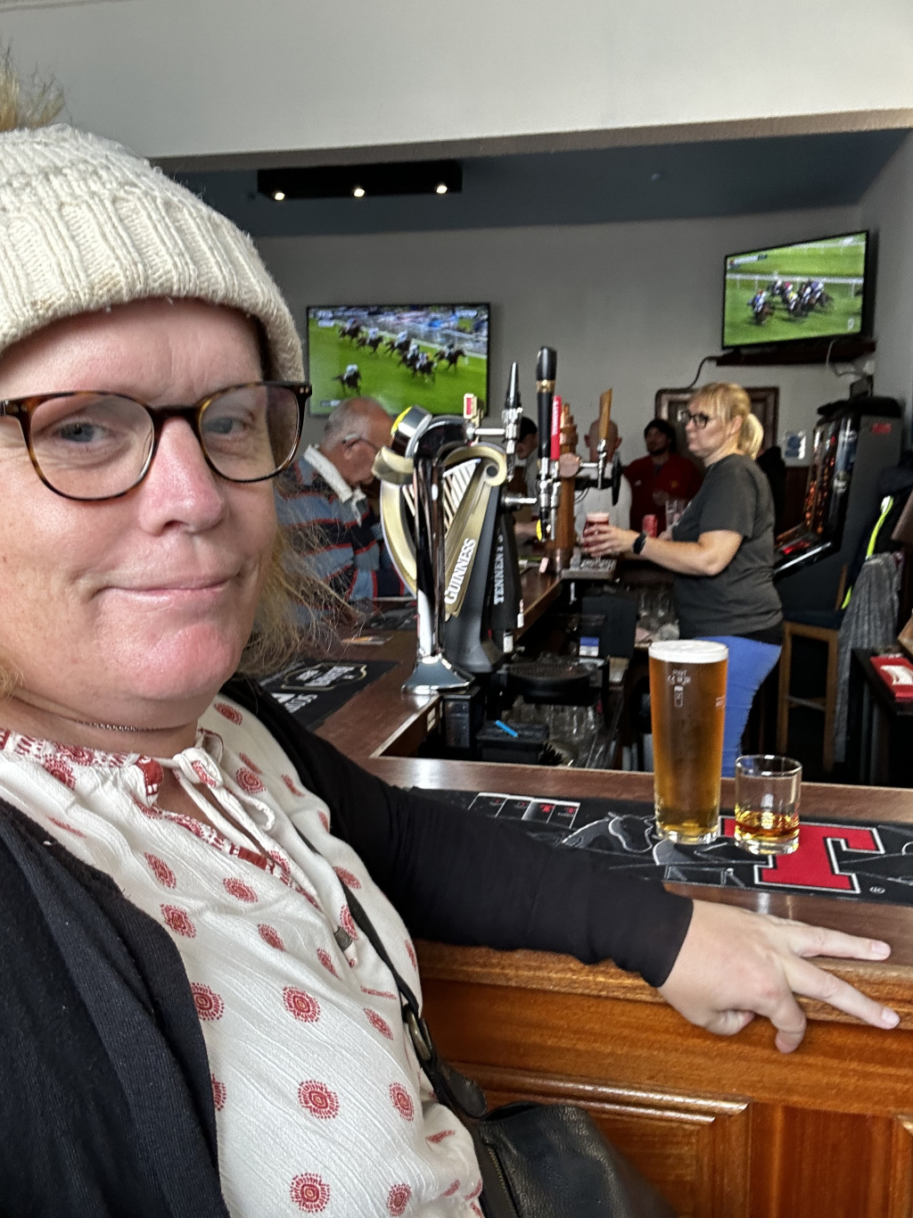 Leonie  sitting at a pub bar. She's wearing glasses and a knit cap. Two television screens in the background show horse races. A pint of beer and a shot glass sit on the bar in front of her. Other patrons are visible at the bar, as is a bartender. The overall atmosphere appears casual and relaxed.