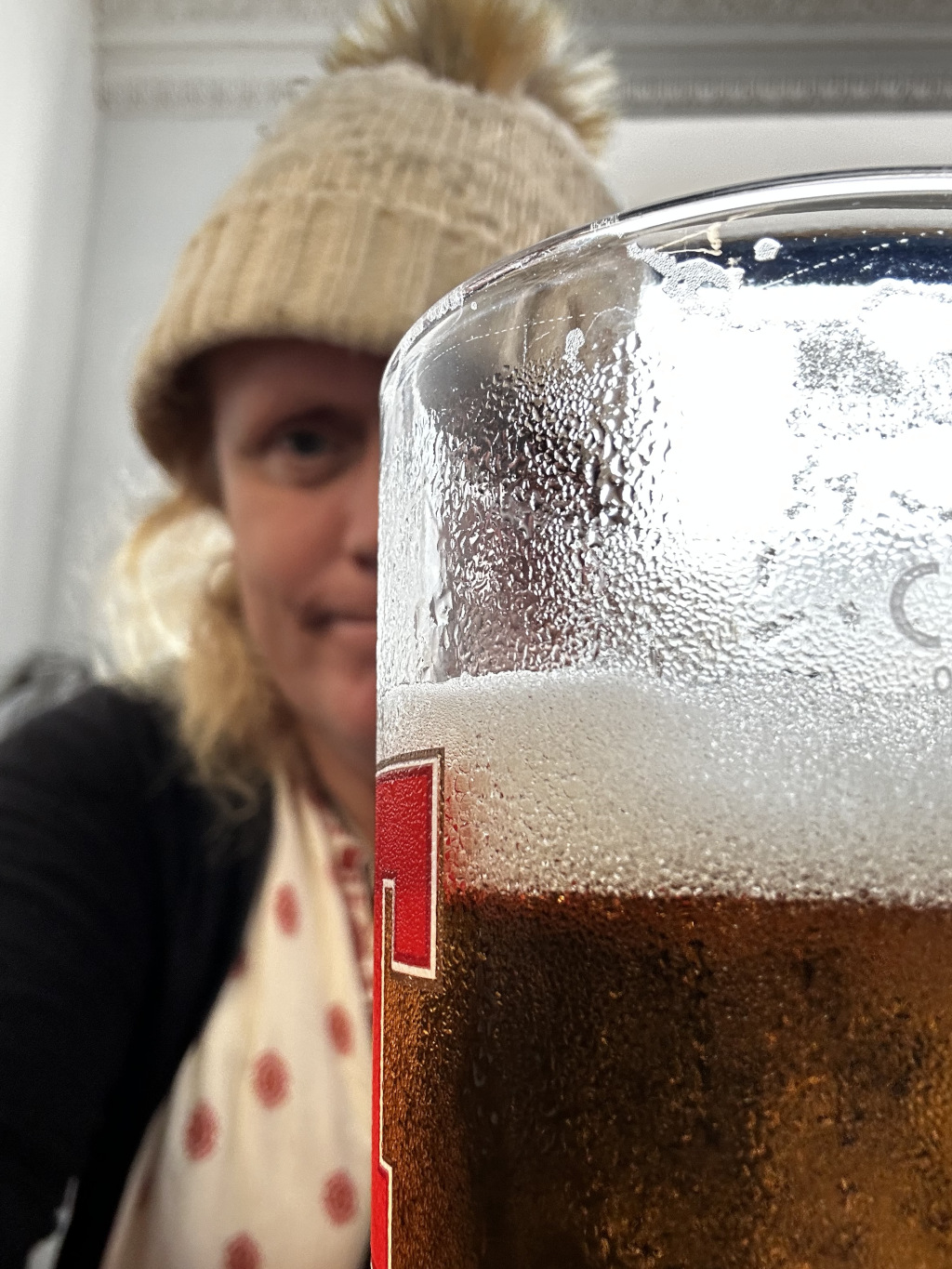 Close-up of a glass of beer, with Leonie in a knit hat blurred in the background. The beer glass is cold, as evidenced by the condensation on its surface. She appears to be taking a selfie or a photo from her perspective. The focus is strongly on the beer, making Leonie a secondary element of the composition.
