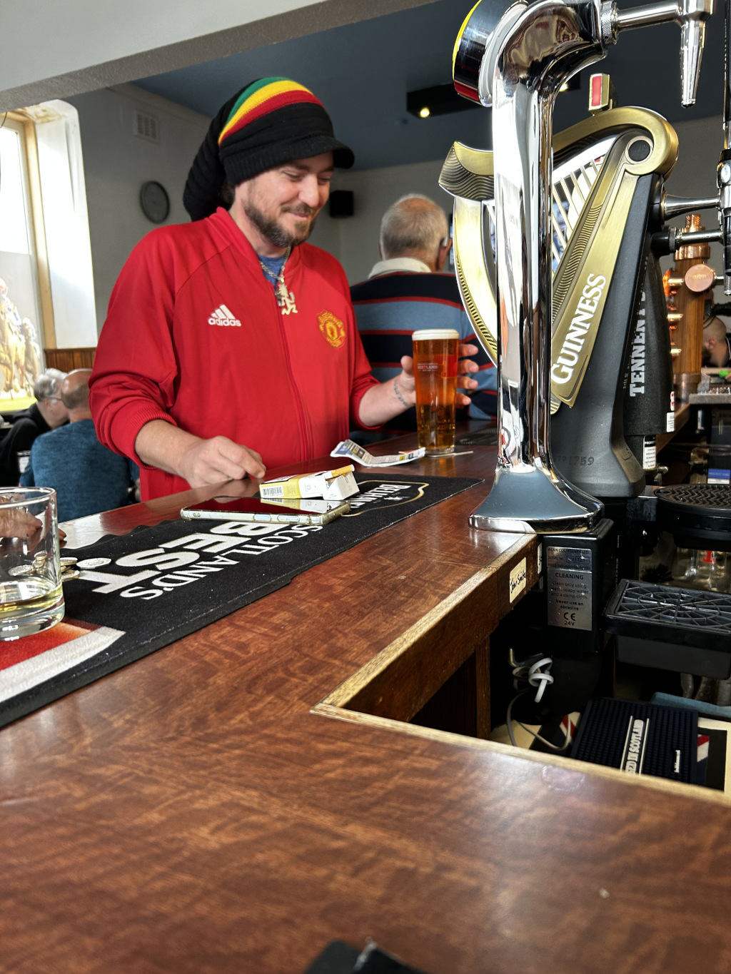 Man with a vibrant red Adidas Manchester United tracksuit, a black knit cap adorned with green, yellow, and red stripes, and a slightly scruffy beard. He's light-skinned with a jovial expression, leaning slightly forward on a dark wood bar top. His hands are occupied; one holds a pack of cigarettes, the other a nearly empty pint glass of amber liquid, likely beer. He seems relaxed and casually at ease. Behind him, partially visible, is a man wearing a blue and red patterned shirt, seemingly engrossed in something else. To the far left, a sliver of another person’s head and shoulder is seen. Dominating the right side is a gleaming, polished chrome and gold Guinness beer tap, reflecting light and showing its brand prominently. It's a prominent vertical element, contrasting with the horizontal lines of the bar.