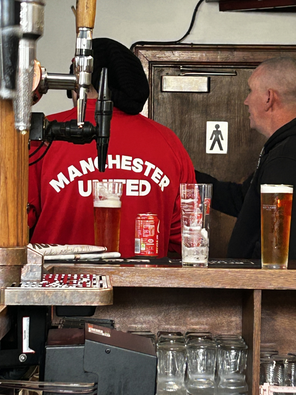 Man wearing a vibrant red Manchester United sweatshirt, the words MANCHESTER UNITED boldly arched across the back. They are positioned slightly angled, with their back to the camera, their head is obscured by a black beanie or cap. They appear to be at the bar, seemingly about to pour a drink from the beer tap just out of frame. The scene is set in a traditional-style pub. The background includes a dark wooden door with a simple, white restroom sign (a stick figure) on it. The walls appear dark, and the lighting is low, creating a somewhat moody atmosphere.