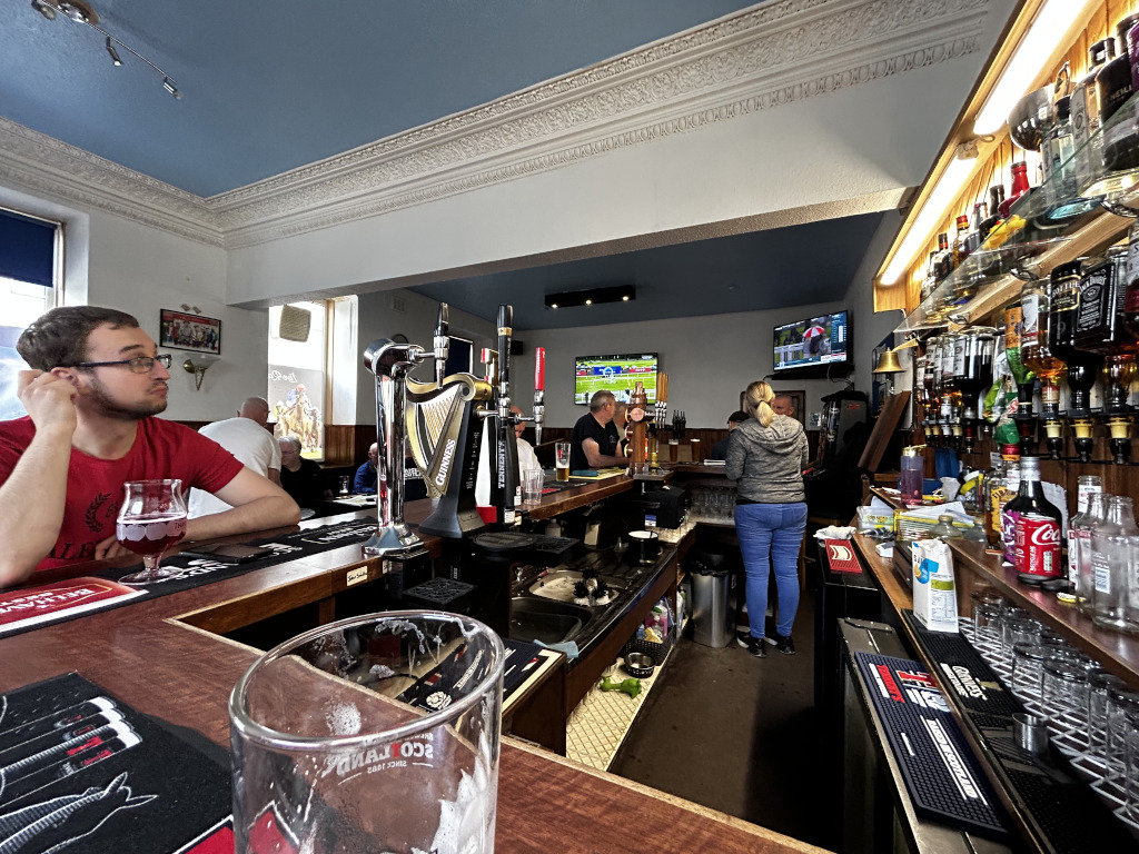 Dominating the foreground is a slightly blurry, empty pint glass resting on a dark wood bar top. A dark brown, textured bar mat with indistinct designs sits just behind it. A portion of a coaster with what appears to be a pub logo is visible on the bar. The main focus is a man in his late twenties or early thirties with glasses. He has light skin and reddish-brown hair, and wears a dark-red t-shirt with a subtle design. He’s seated at the bar, his left hand resting on his head, looking to the right, exhibiting a relaxed but slightly pensive expression. Next to him is a half-full stemmed glass with a reddish-amber liquid, likely alcoholic, suggesting a relaxed pub setting. Behind him, other patrons are sparsely seated at the bar, partially obscured. Beyond them is the bar itself, a long, dark wood surface with a highly polished sheen. A gleaming Guinness tap system is prominently featured on the bar, adding depth to the image. The bar extends across the image, disappearing into the background. A woman in a dark jacket and blue jeans stands towards the middle background, facing the bar. Her posture suggests she’s waiting or having a conversation.