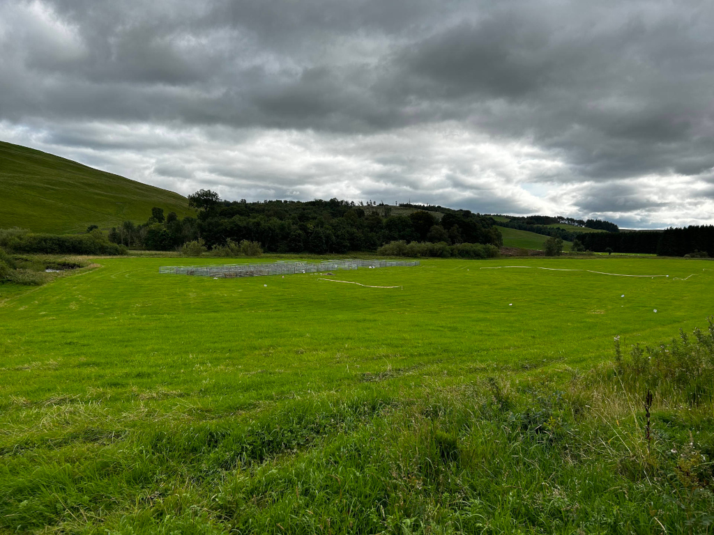 Wide, expansive green field under a cloudy sky. In the mid-ground, there is a series of what appear to be temporary livestock pens or enclosures made of metal fencing. Beyond the field, there's a line of trees and rolling hills. T