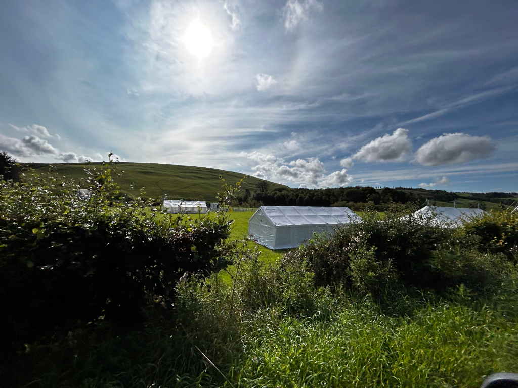 Sunny landscape with several white tents pitched in a grassy field. The tents appear to be temporary structures, possibly for camping or an event. Rolling green hills and a partly cloudy blue sky form the backdrop. The scene is peaceful and idyllic.