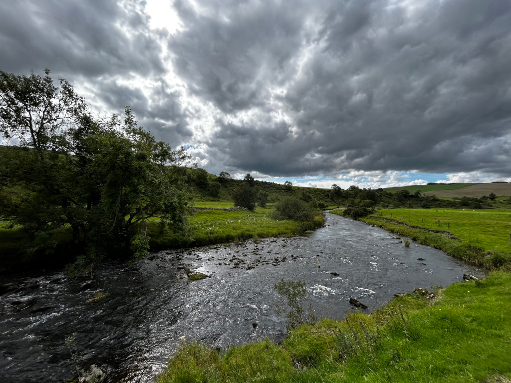 Tranquil river scene under a dramatic, heavily clouded sky. A gently flowing river meanders through lush green pastures, bordered by verdant banks and a few trees. The sky is a mix of dark, brooding clouds and lighter areas, suggesting an impending change in weather. 