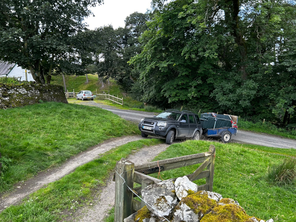 Rural landscape with a dirt road leading to a house.  A dark-colored SUV towing a small trailer loaded with items (possibly luggage or camping gear) is parked on the road.  Another smaller, light-colored car is parked further up the road, near a stone wall. The setting is lush and green, with mature trees lining the road. A wooden gate is in the foreground. 