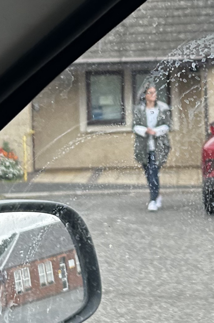 Taken from inside a car, shows a blurry view of a woman walking in front of a building. The woman is wearing a light-colored jacket and dark pants. The building appears to be light-coloured with windows. The car's windshield is dirty, obscuring the view. The reflection of another building shows in the car's side mirror. 