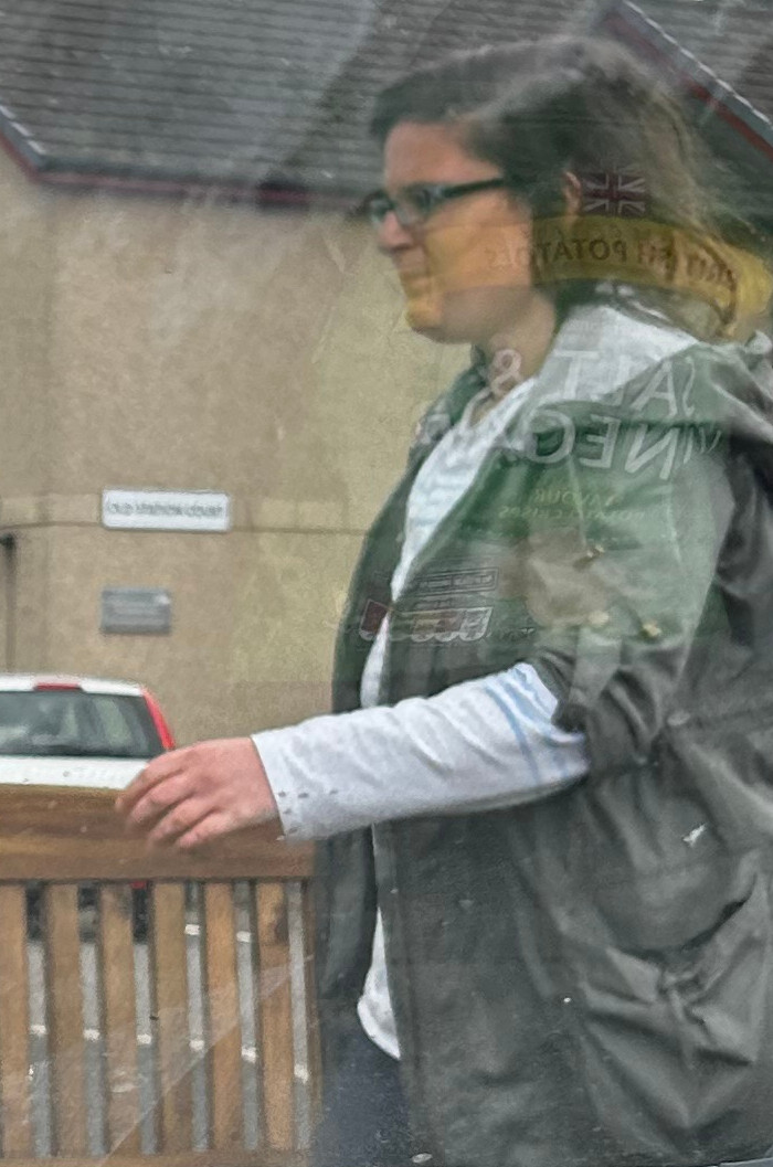 Person, seemingly a woman, wearing glasses and a dark-coloured jacket. She appears to be walking past a wooden bench, outside of a building. The image is slightly blurry and out of focus, possibly taken through a window or some type of protective covering. The background shows a light-coloured building and the partial view of a car. 