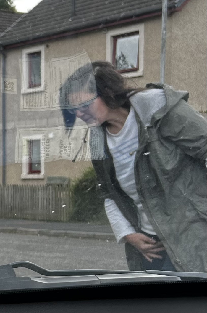 Woman viewed through a car windshield. She is wearing a green jacket and appears to be looking down, possibly at something on the ground. The windshield is somewhat dirty or smeared, and there are several pieces of paper stuck to the outside of it, obscuring the view. The background shows a residential building.