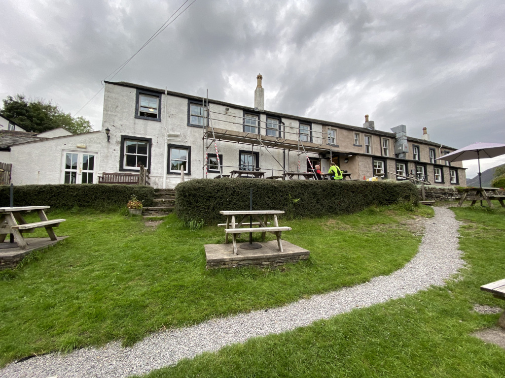 Two-story building, possibly a pub or inn, undergoing renovations. Scaffolding is visible on the front of the building. The building is white with dark window frames. The landscaping includes a grassy area with picnic tables and a gravel pathway. Two people are visible on the patio area in front of the building. The overall atmosphere is somewhat overcast.