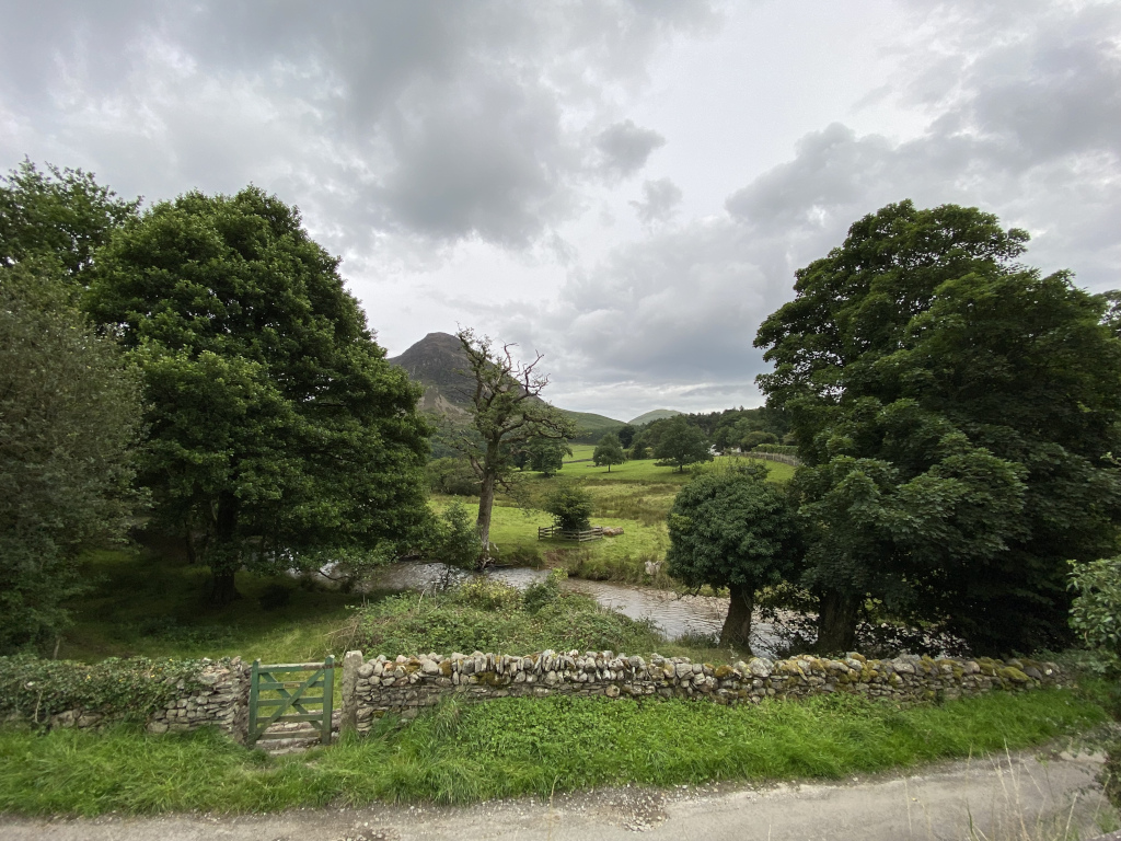 Tranquil pastoral scene, likely in a rural area of the UK.  A small stream meanders through a grassy field, bordered by lush green trees and a low stone wall with a small green gate. In the background, a softly rounded hill rises under a sky filled with fluffy, grey clouds. 