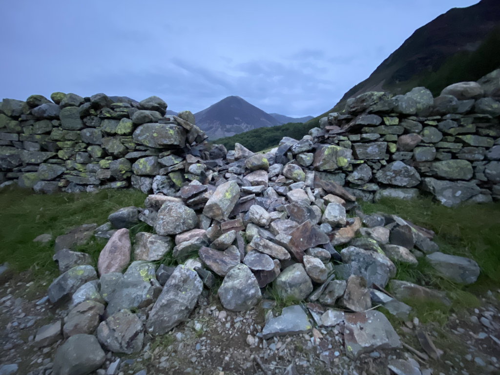 Pile of rocks in the foreground, forming a gap or break in two low stone walls that extend to either side. The walls appear to be old and weathered, with lichen or moss growing on some of the stones. In the background, there are mountains under a partly cloudy sky.