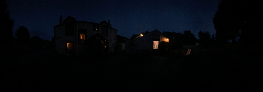 Two buildings at night, mostly shrouded in darkness. A larger building, possibly a house or inn, is on the left, with a few dimly lit windows visible. To the right, a smaller structure has a single brightly lit window. The surrounding area is dark, with silhouettes of trees and bushes suggesting a rural or secluded setting. The overall mood is one of quiet solitude and mystery.