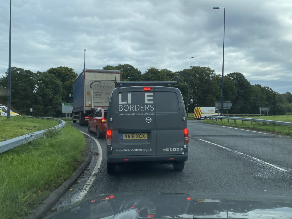 Traffic jam on a road. A dark-grey van with the words LIE BORDERS prominently displayed on its side is in the foreground. Behind it are other vehicles including a red car and a large lorry. The setting appears to be a rural road with trees lining the sides. The overall impression is of a slightly congested road scene, with the van's advertisement being a key focal point.