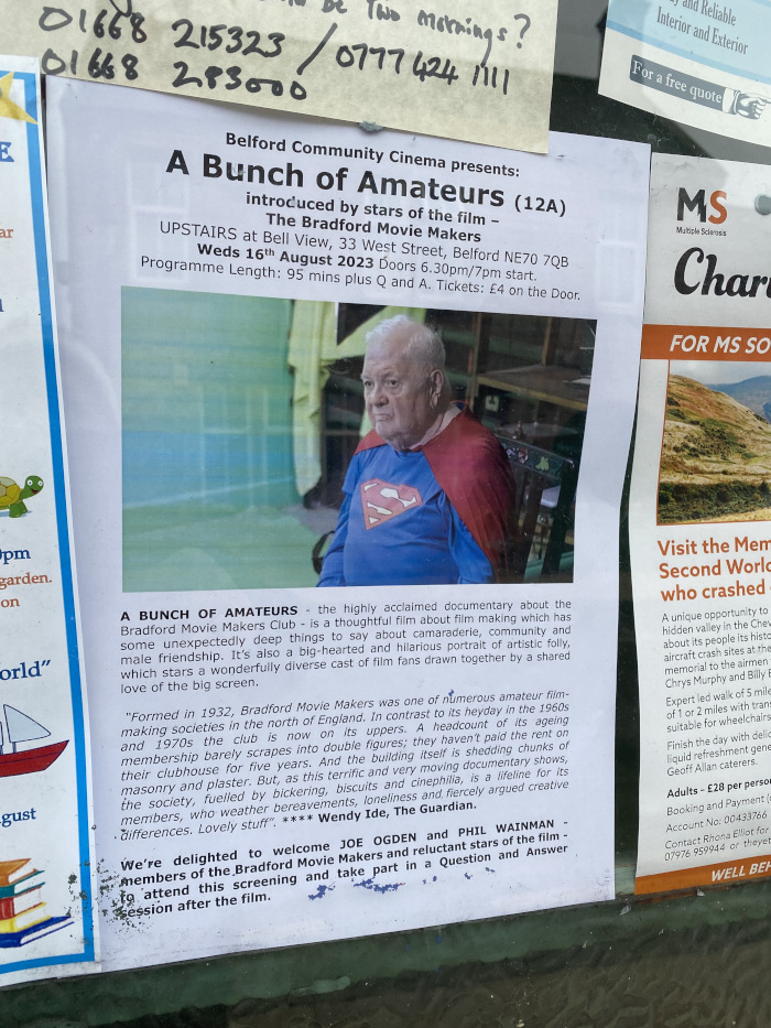 Poster advertising a showing of the documentary A Bunch of Amateurs at the Belford Community Cinema. The poster features a photo of an older man dressed as Superman, one of the film's subjects. The text describes the film as a thoughtful and humorous documentary about the Bradford Movie Makers Club, focusing on their camaraderie, community, and the challenges faced by an aging group of amateur filmmakers. The poster also includes details about the screening time, date, location, and ticket price. Other unrelated posters are visible in the background.