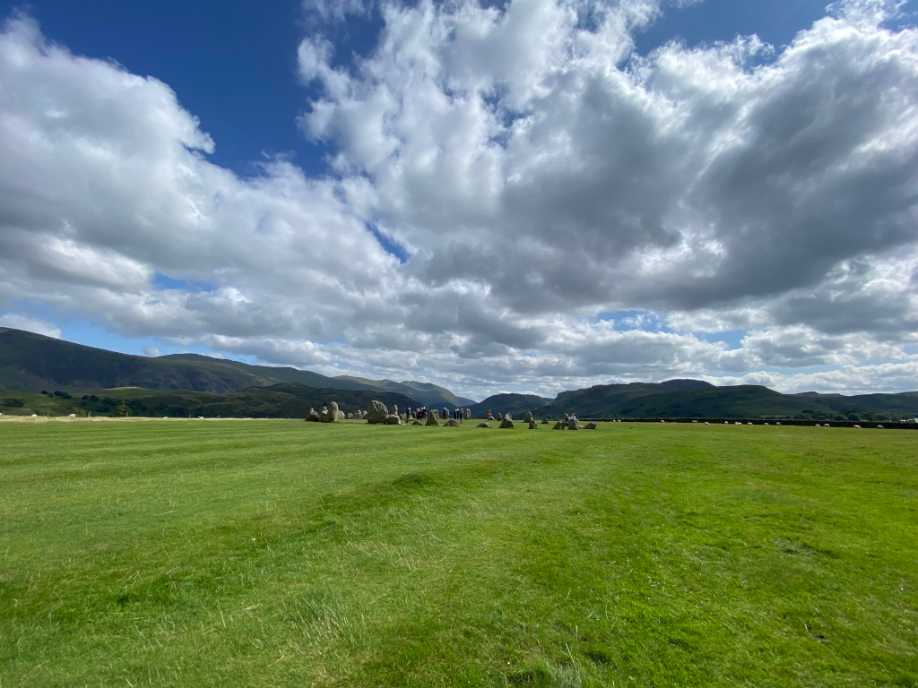 Vast, verdant field stretching towards a low mountain range under a partly cloudy sky. In the middle ground, a line of large stones, possibly a stone circle or similar ancient structure, is visible. A small number of people are present near the stones, providing a sense of scale. The overall scene is peaceful and evokes a sense of wide-open space and natural beauty.