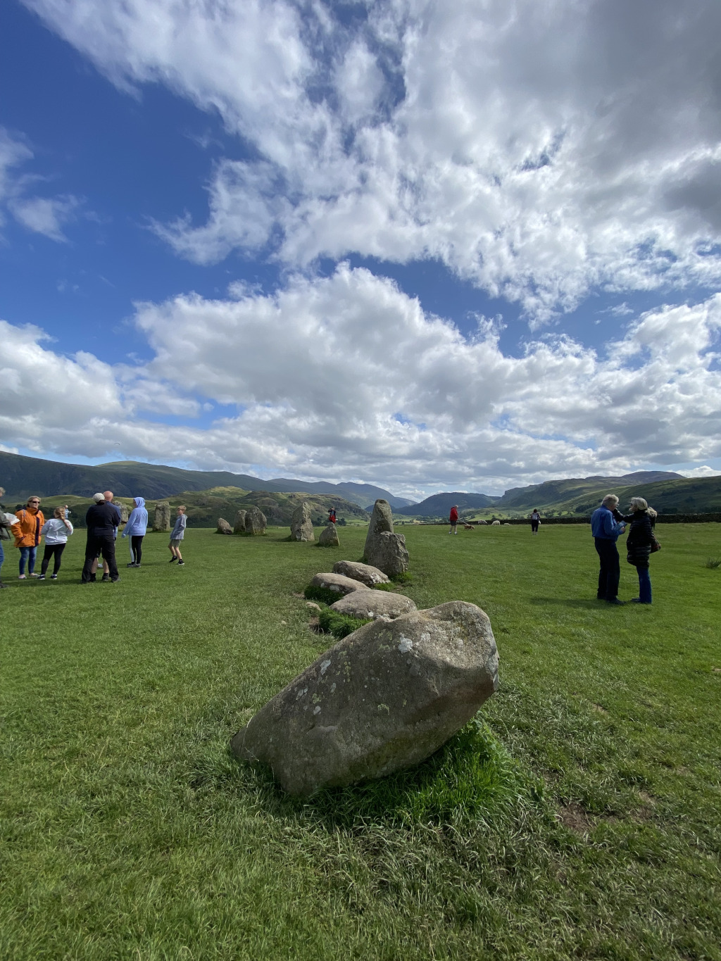 partially-fallen stone circle in a grassy field, under a mostly sunny sky with fluffy cumulus clouds. Several people are visiting the site, observing the stones from a short distance. Rolling hills are visible in the background, under a bright blue sky. The overall impression is one of a peaceful and historic setting.