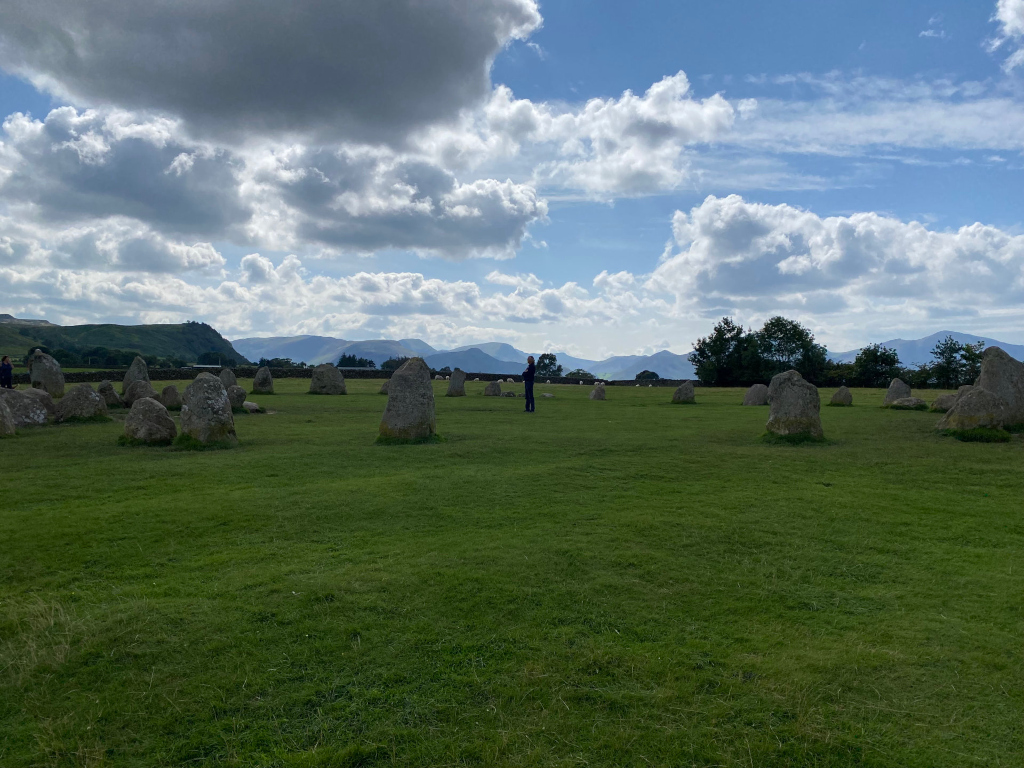 Stone circle in a grassy field, with a backdrop of rolling hills and a partly cloudy sky. A few people are visible in the distance, giving a sense of scale to the ancient stones. The overall mood is peaceful and evocative of a historical or possibly spiritual site.
